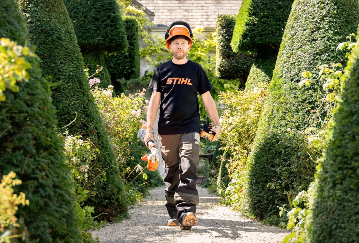 Gardener Andrew Wain in the middle of a row of topiary trees in the shape of a cone. In his right hand he carries a pruner, in his left a battery pack.