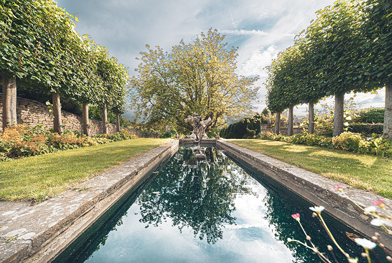 An elongated, square water basin, framed by strips of lawn and espaliered lime trees.
