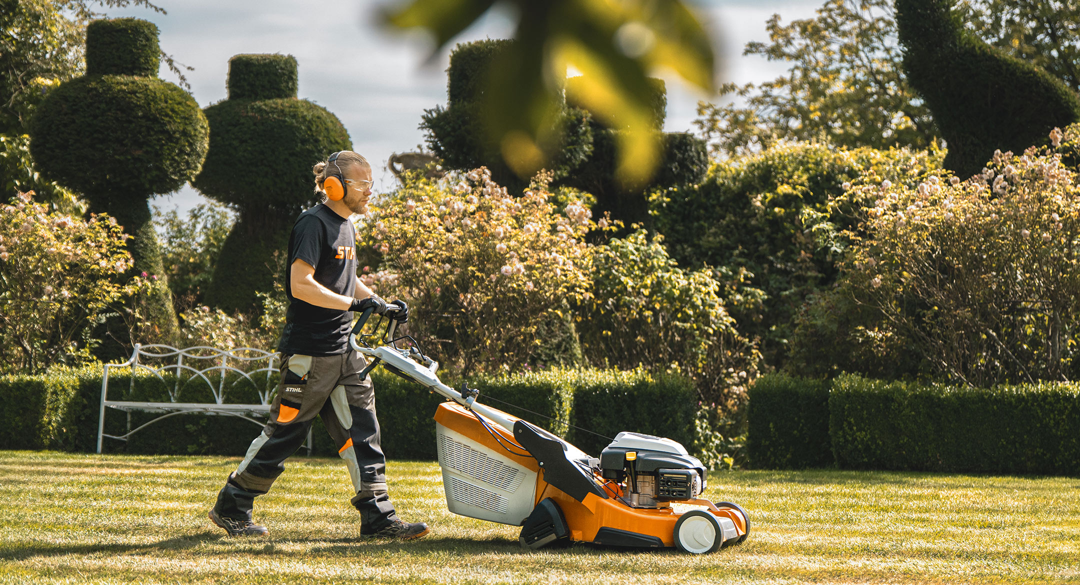 Andy Wain pushes a lawnmower across a green area. In the background are the chess pieces made from yew trees.
