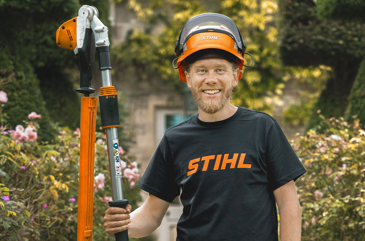 Andy Wain stands smiling in front of a rose bed, a pole pruner in his right hand