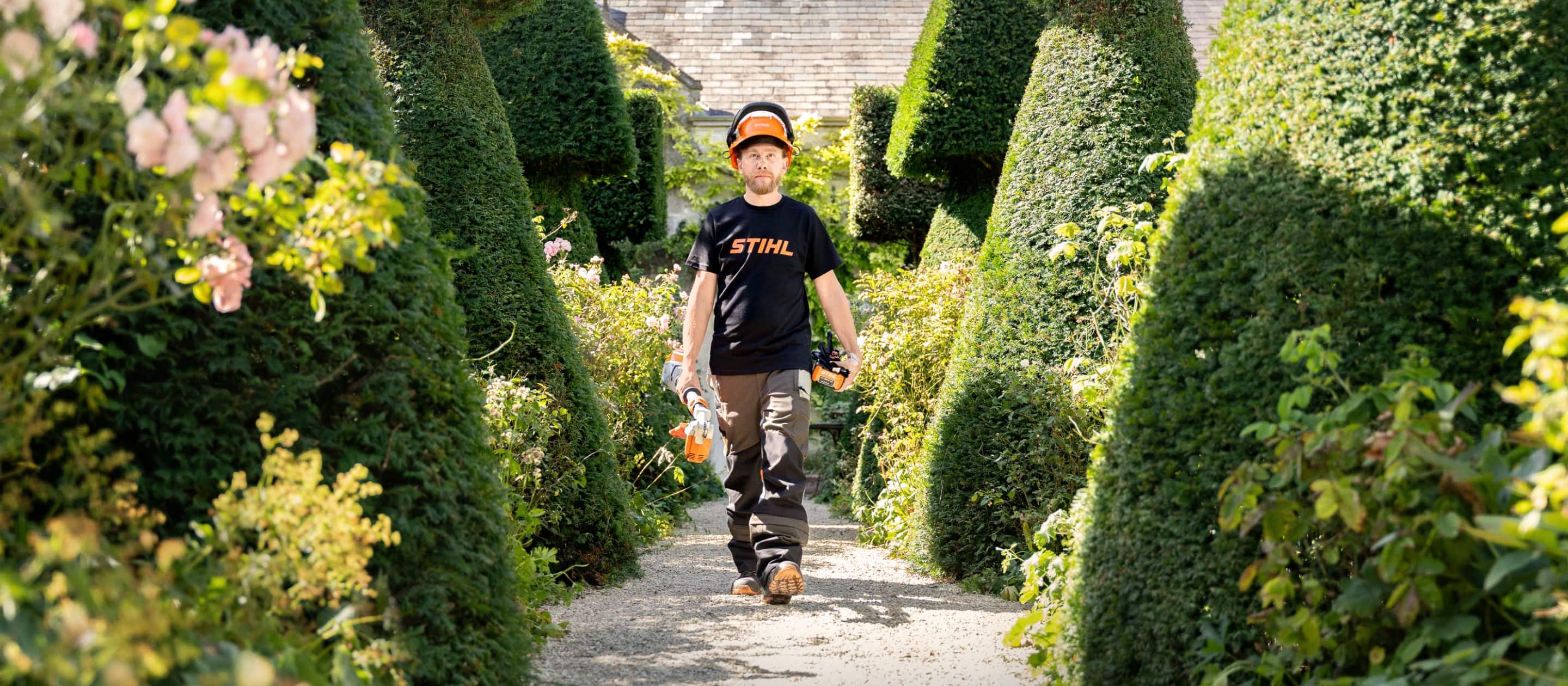 Gardener Andrew Wain in the middle of a row of topiary trees in the shape of a cone. In his right hand he carries a pruner, in his left hand a battery pack