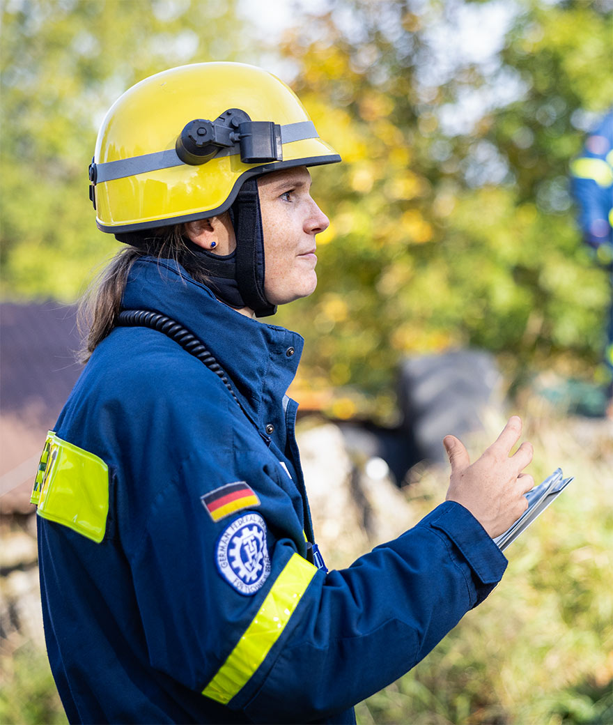 Anna Mirl in profile, in blue THW uniform and yellow hard hat, a clipboard in her hand.